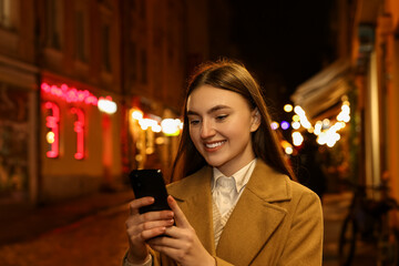 Smiling woman using smartphone on night city street. Space for text