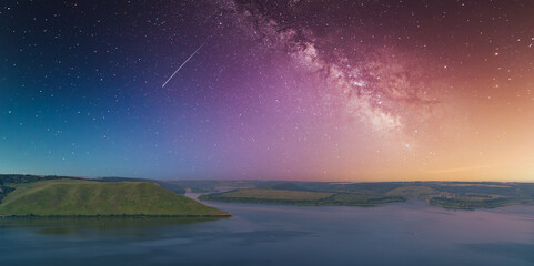 Cosmic night sky over a calm river and green hills, with the Milky Way and a shooting star visible