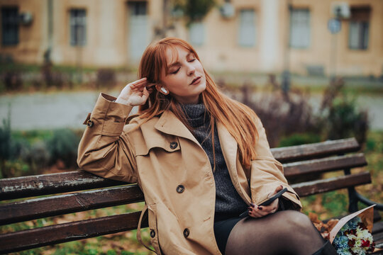A young trendy woman is sitting on a bench in park and enjoying music.