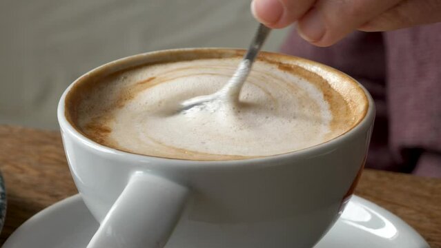 Hand stirring Cappuccino coffee in a white ceramic cup with a silver spoon on a rustic wooden tabletop, close-up shot.