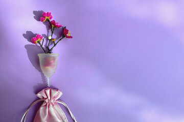 Menstrual cup with red flowers and pink silk bag, flat lay on purple background, overhead shot....