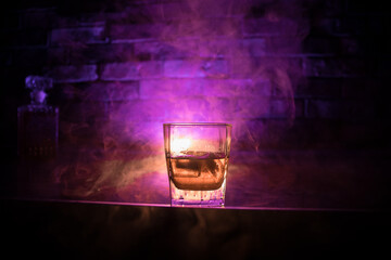 Whiskey in fire concept. Glass of whiskey and ice on wooden surface with color light and fog on...