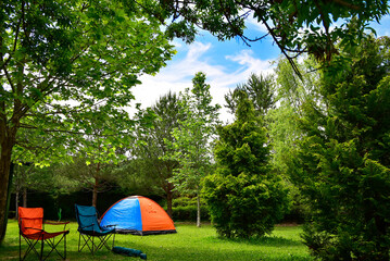 Orange camping tent and camping chairs next to it among the trees in the green forest