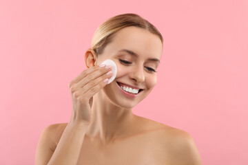Smiling woman removing makeup with cotton pad on pink background - obrazy, fototapety, plakaty