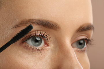 Woman applying mascara onto eyelashes against light brown background, closeup