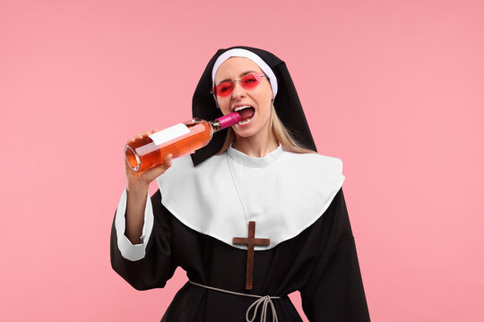 Woman in nun habit and sunglasses holding bottle of wine on pink background