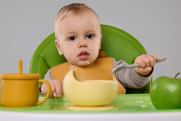 Cute little baby eating healthy food in high chair on gray background