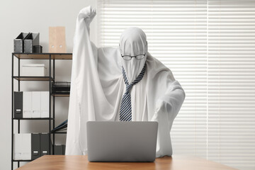 Overworked ghost. Man covered with white sheet using laptop at wooden table in office