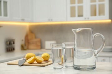 Jug, glasses with clear water and lemons on white table in kitchen