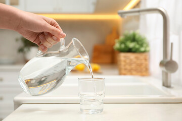 Woman pouring water from jug into glass at white table in kitchen, closeup. Space for text