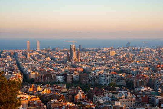 Panoramic view of Barcelona at sunrise or sunset with golden light over the cityscape, featuring the Sagrada Familia and prominent skyscrapers like Torre Glories and Hotel Arts.