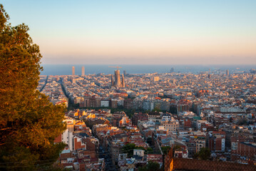 Panoramic view of Barcelona at sunrise or sunset featuring the Sagrada Familia and Eixample's grid pattern. Warm hues highlight the Mediterranean architecture and city's proximity to the sea.