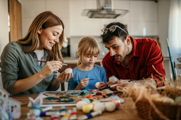 A joyful family of three gathered around a dining table, enthusiastically painting vibrant Easter...