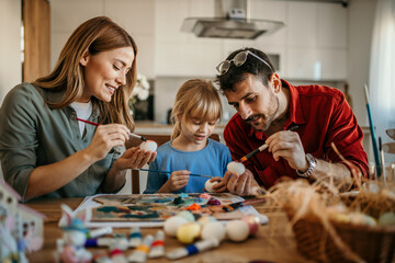 A joyful family of three gathered around a dining table, enthusiastically painting vibrant Easter...