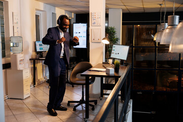 Happy manager listening music during work break, dancing in startup office after finishing marketing strategy. African american businessman enjoying having fun late at night in startup office