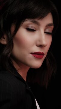 A beautiful brunette woman in a black business suit and red lips posing in the studio on a dark background. Beauty face. Vertical video
