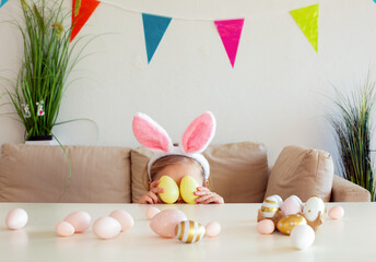 Blond child girl wearing bunny ears on Easter day play with Easter eggs at the table at home
