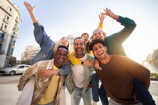 Portrait excited multi-ethnic group happy young friends piggyback enjoying great time outdoors and having fun together. Cheerful men carrying girlfriends on back. Looking at camera on vacation