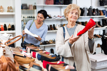 Elderly woman selecting new footwear in shoe store. Asian woman seller helping her.