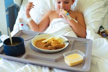 Cute European 7-8 year old boy eats hospital food while lying in the bed of a personal room with a catheter in his hand - 728134756
