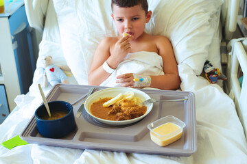 Cute European 7-8 year old boy eats hospital food while lying in the bed of a personal room with a catheter in his hand - 728134768
