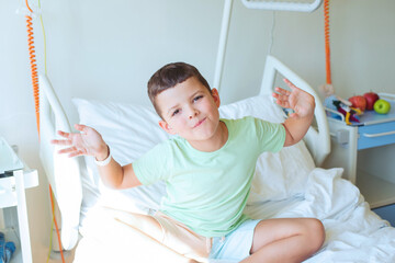 Portrait of a smiling European seven year old boy in a light green T-shirt sitting on the bed in a hospital room - 728134773