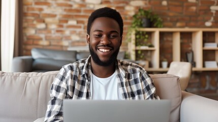 Fototapeta premium Smiling young black man working or communicating online, sitting on couch with laptop at home. Cheerful millennial guy freelancing, chatting on internet. Modern technologies in everyday life concept