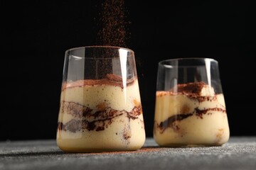 Adding cocoa powder into glass with delicious tiramisu at grey textured table, closeup