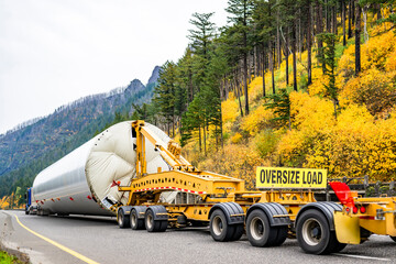 Blue big rig semi truck transporting oversized part of the wind generator support pole using an additional heavy duty trolley with six axles and oversize load sign driving on the autumn highway road