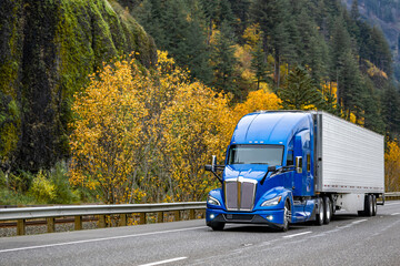 Bonnet blue big rig semi truck tractor carry cargo in refrigerated semi trailer driving on the autumn highway road with rock mountain range on the side - obrazy, fototapety, plakaty
