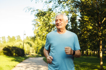 Naklejka na ściany i meble An elderly man in a blue T-shirt is jogging through a summer park.