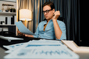 Smart businessman holding paperwork report paragraph dynamic database, looking at pc screen with analyzing global target evaluation value profit company at modern home office at night time. Pecuniary.