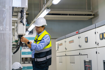 Professional Asian man engineer in safety uniform working at factory server electric control panel room. Industrial technician worker maintenance checking power system at manufacturing plant room.