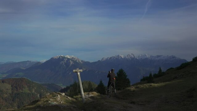 Male cyclist on gravel bike enjoying view and making phono on phone on top of mountain in alps with beautiful view of snowy peaks of mountain range in germany. Sports motivation and inspiration. 