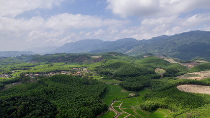 Aerial view of lush green rolling hills and agricultural fields with a small village nestled among the trees, under a cloudy sky background