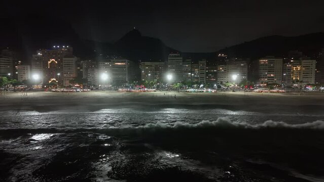 Aerial above breaking waves along Copacabana Beach at night. Oceanfront street and buildings with city lights. Corcovado Hill in background
