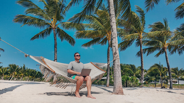 Man with a laptop sitting in a hammock between palm trees on a sunny beach, symbolizing remote work or a workation concept