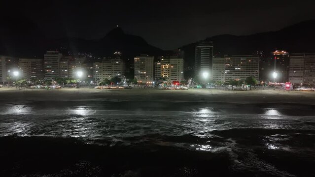 Aerial above breaking waves along Copacabana Beach at night. Oceanfront street and buildings with city lights. Corcovado Hill in background
