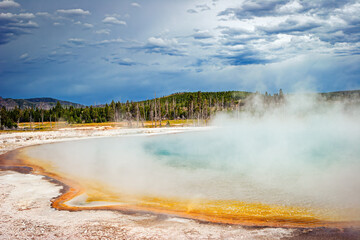 Colorful Yellowstone National Park geyser with hot steam, hillside trees and storm clouds, Wyoming, USA. 