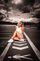 A young blonde woman sits in a parking lot on the markings in the background of a dramatic stormy...