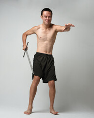 Fototapeta premium Full length portrait of fit handsome asian male model, wearing gym shorts and shirtless. Holding sword weapons whilst in warrior training action poses, isolated on white studio background.