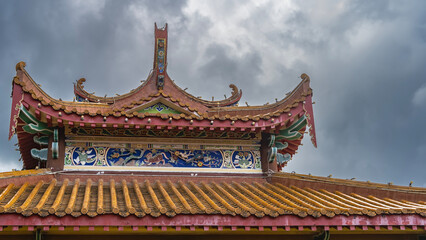 Fototapeta na wymiar Details of the architecture of the Chinese Thean Hou Temple. The tiled roof with curved edges is decorated with carved ornaments and drawings. The background is a cloudy sky. Malaysia. Kuala Lumpur.