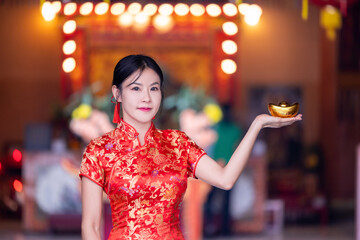 Beautiful Asian woman wearing a traditional red cheongsam on Chinese New Year. Young woman hand holding red envelope or Ang pao. Asian woman holding gold ingot in Chinese Buddhist temple. .
