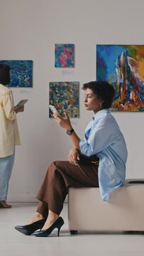 Full vertical shot of young African American female visitor in casual clothes and stiletto heel shoes sitting on couch in art gallery at modern art exhibition, using smartphone and resting