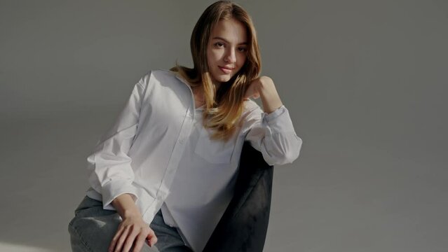 Beautiful young woman in white shirt sitting on chair and posing on camera during studio shooting. Charming female model smile and looking at camera