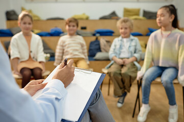 Close up of teacher or therapist holding clipboard and taking notes while working with group of...