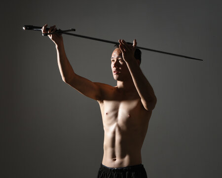 portrait of fit handsome asian male model, wearing gym shorts and shirtless. Holding sword weapons  whilst in warrior training action poses, isolated on moody dark studio background with silhouette