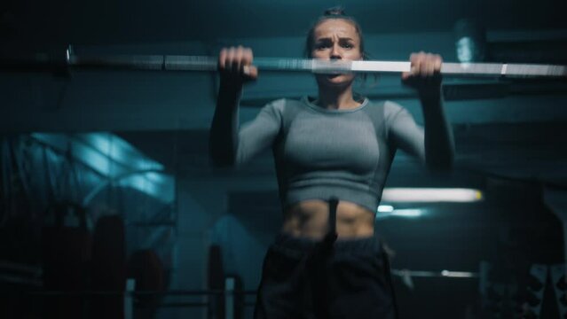 Strong woman with athletic body wearing sportswear lifts and does vigorous exercises with empty barbell in dark gym with LED lighting. Female athlete trains before sport competition. CrossFit workout.