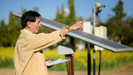 Smiling senior man holding clipboard standing beside solar panel at the cornfield. Renewable energy...