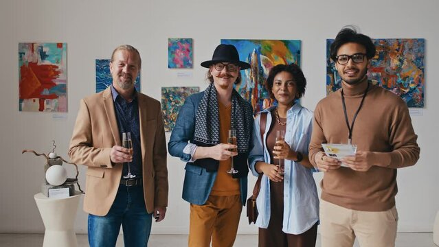 Medium zoom portrait shot of diverse contemporary art gallery team standing in front of paintings at exhibition opening, holding champagne glasses, chatting, then looking at camera and smiling
